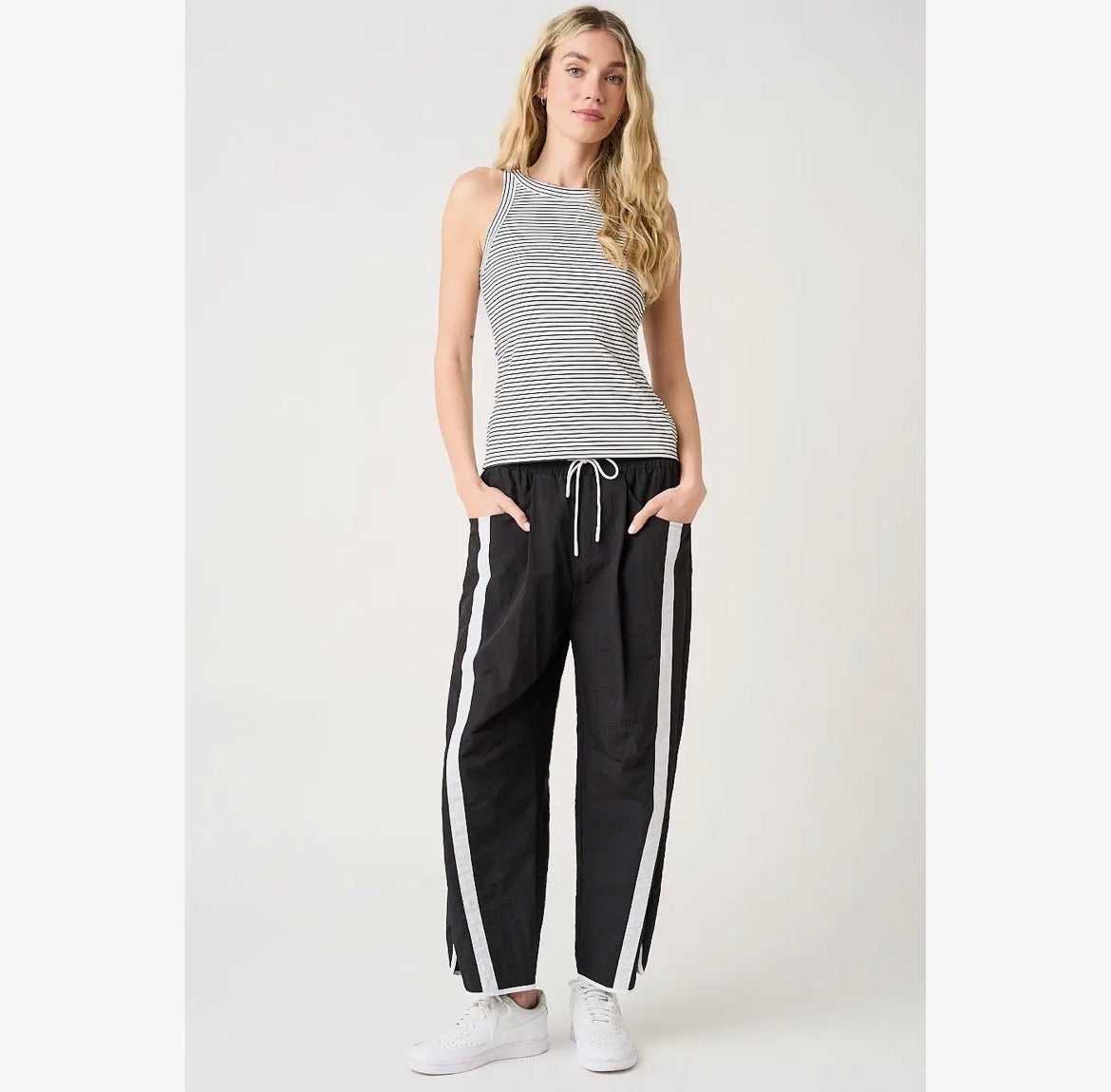 A woman modeling a sleeveless gray and white striped tank top with black wide-leg sweatpants featuring white side stripes, paired with white sneakers, against a light gray studio background.