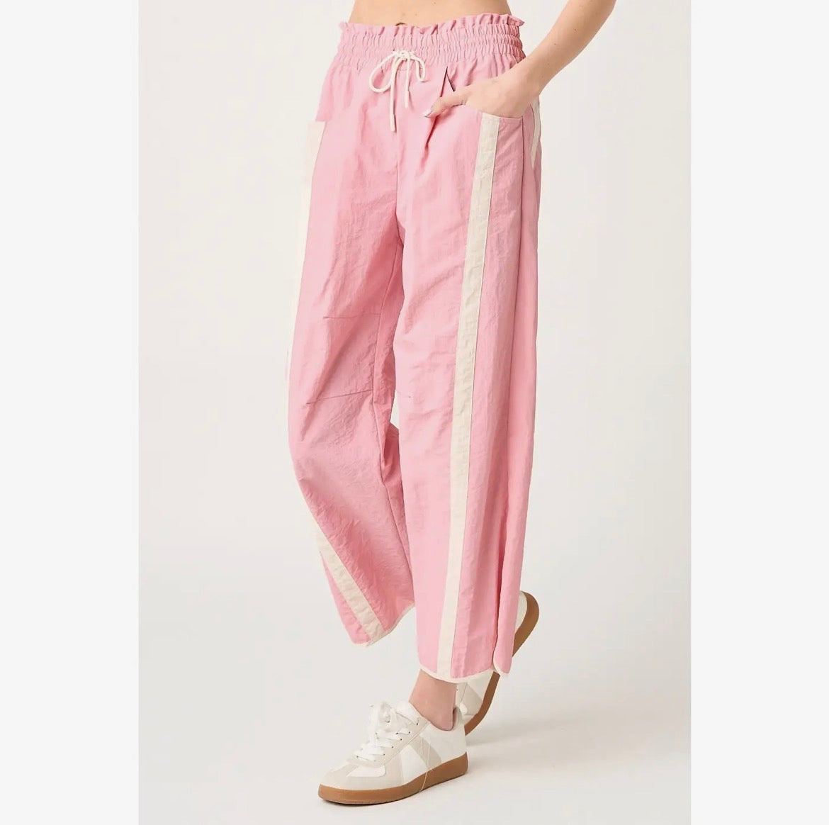 A person wearing loose pink pants with a cream drawstring waist and vertical cream side stripes, paired with white sneakers, photographed against a plain light background.