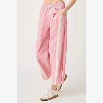 A person wearing loose pink pants with a cream drawstring waist and vertical cream side stripes, paired with white sneakers, photographed against a plain light background.