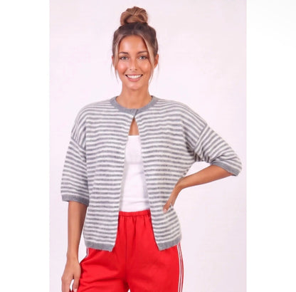 A woman modeling a gray and white striped knit cardigan with a keyhole cutout, worn over a white top and red athletic pants, against a light pink studio background.