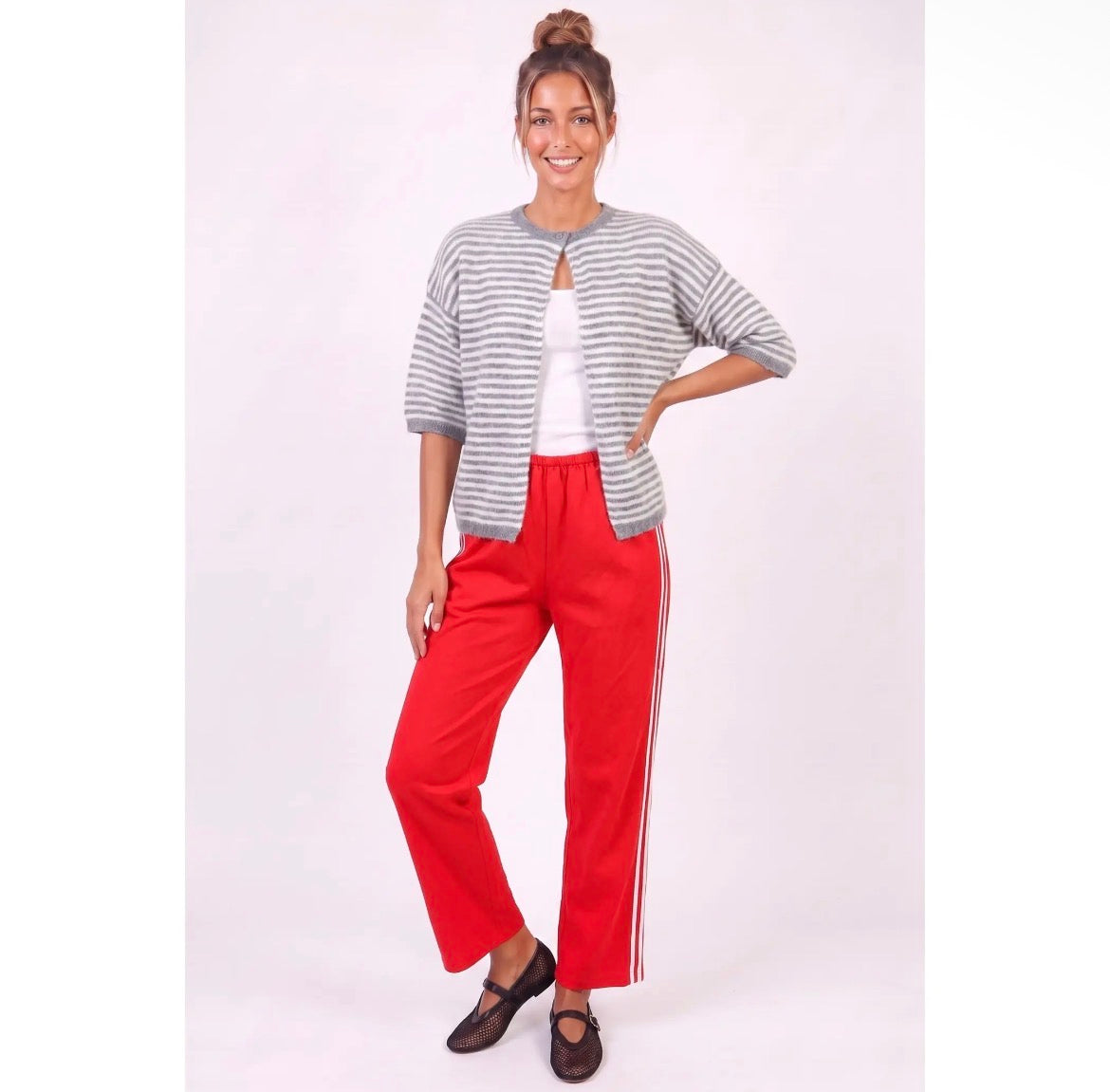 A woman modeling a casual outfit consisting of a gray and white striped short-sleeve cardigan over a white top, paired with bright red wide-leg pants featuring white side stripes, and dark brown flat shoes, against a plain light background.