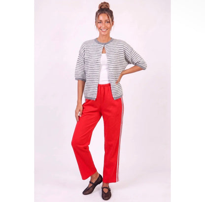 A woman modeling a casual outfit consisting of a gray and white striped short-sleeve cardigan over a white top, paired with bright red wide-leg pants featuring white side stripes, and dark brown flat shoes, against a plain light background.