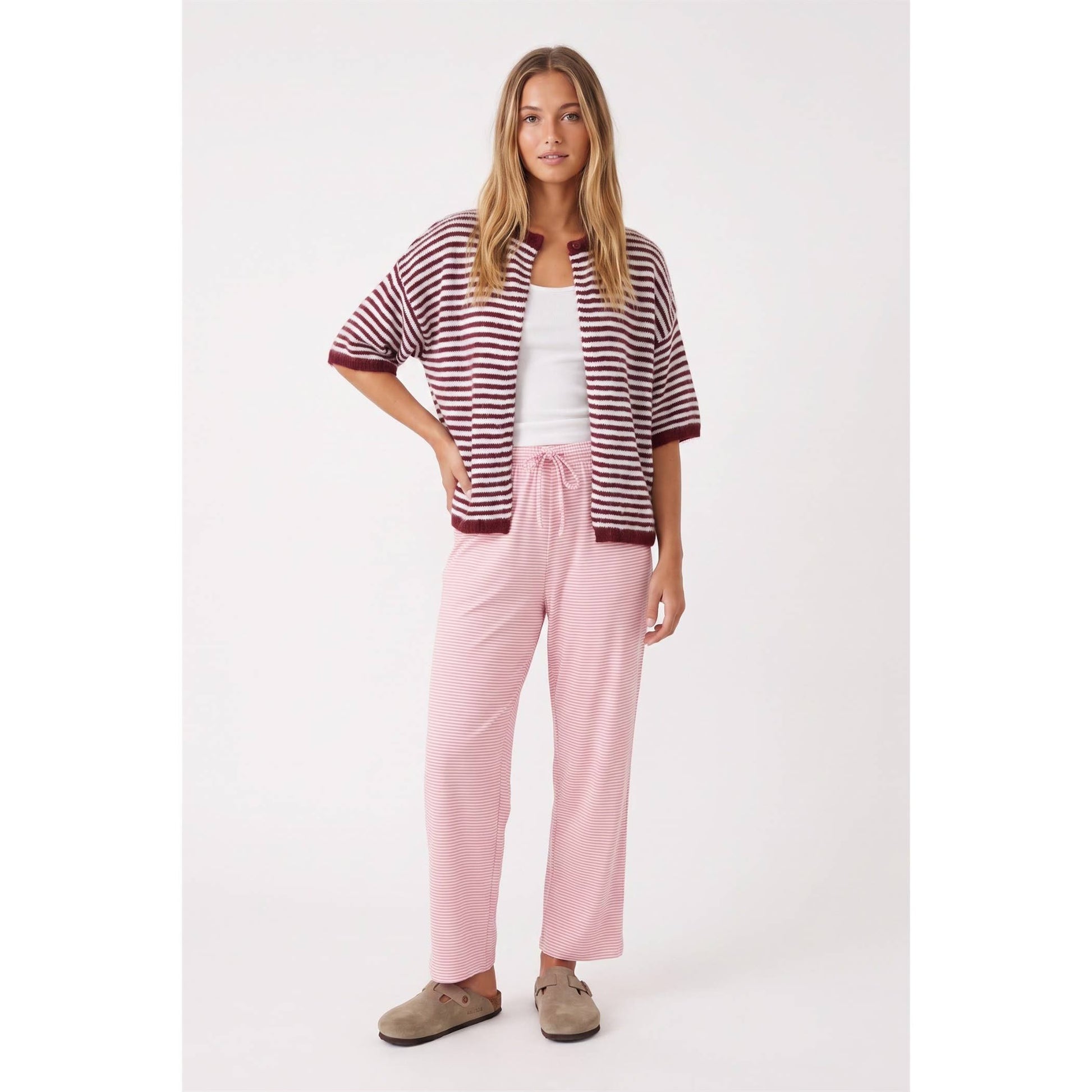 A woman modeling a matching loungewear set consisting of a burgundy and white striped short-sleeve cardigan, a white tank top, light pink drawstring pants, and taupe slip-on shoes, against a plain light gray studio background.