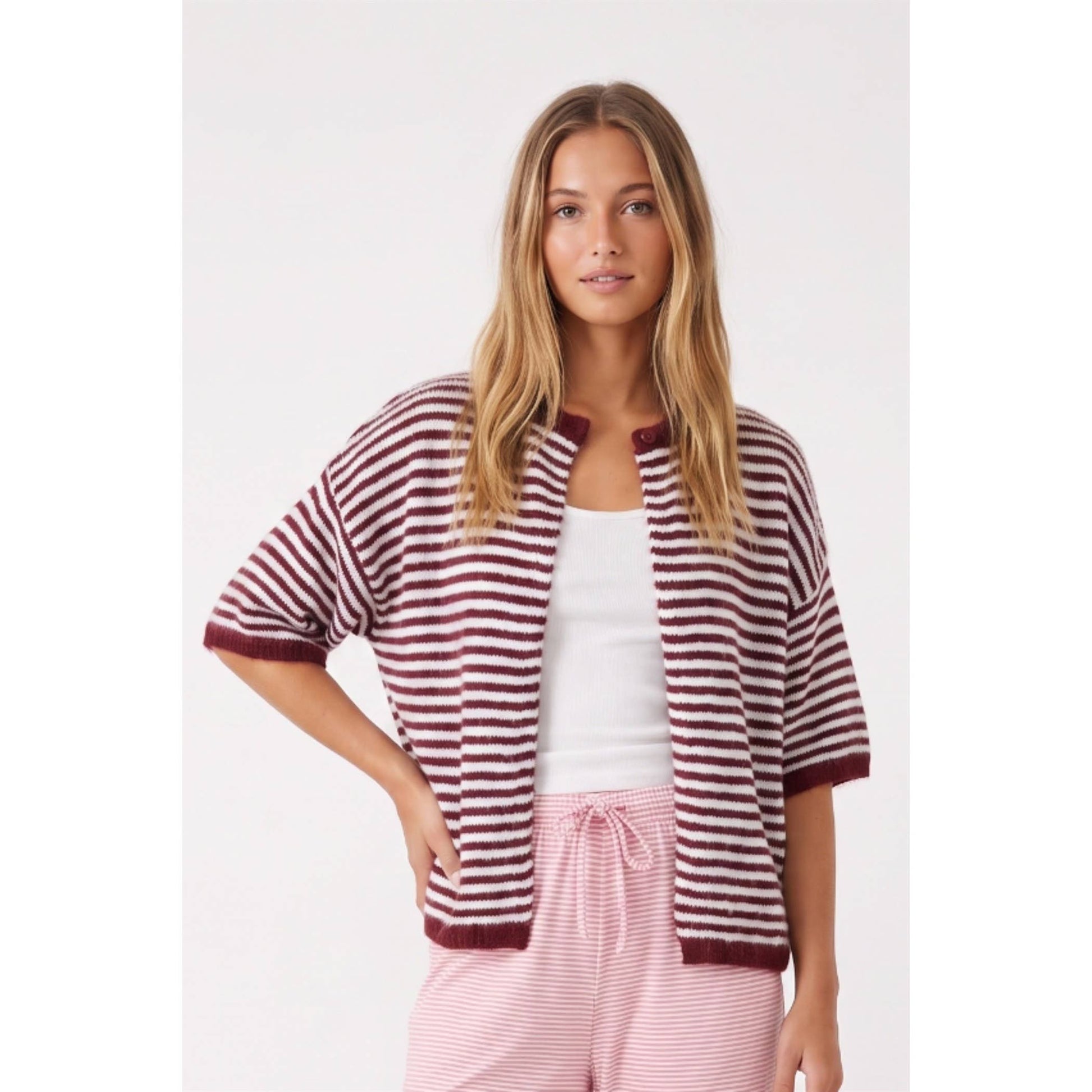 A woman modeling a short-sleeve knit cardigan with horizontal burgundy and off-white stripes, worn open over a white tank top and pink striped pants against a light gray studio background.
