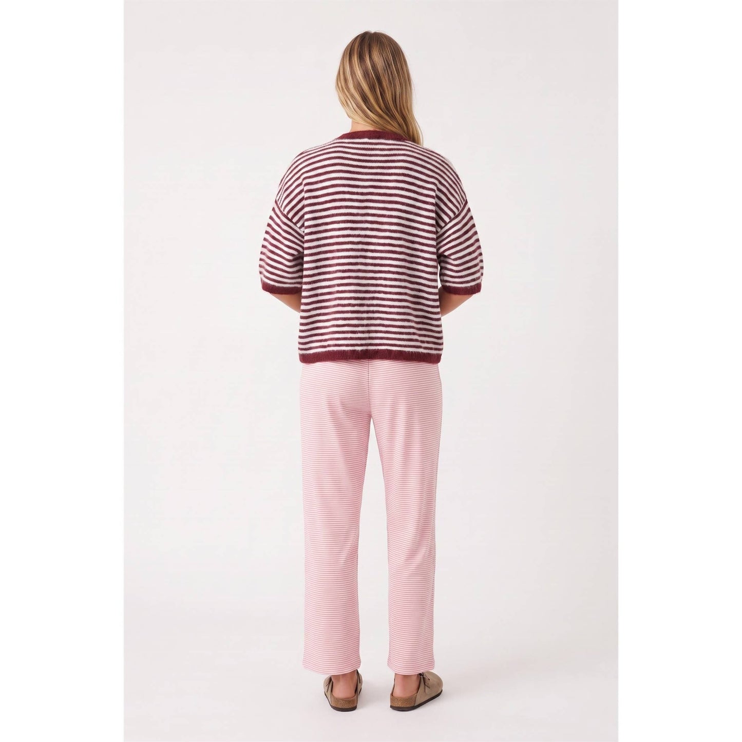A woman viewed from behind wearing a short-sleeve knit top with horizontal burgundy and off-white stripes, paired with light pink pants and brown sandals, against a plain light background.