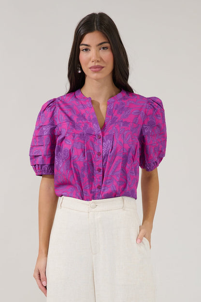 Woman wearing a pink and purple patterned blouse with white pants on a plain background