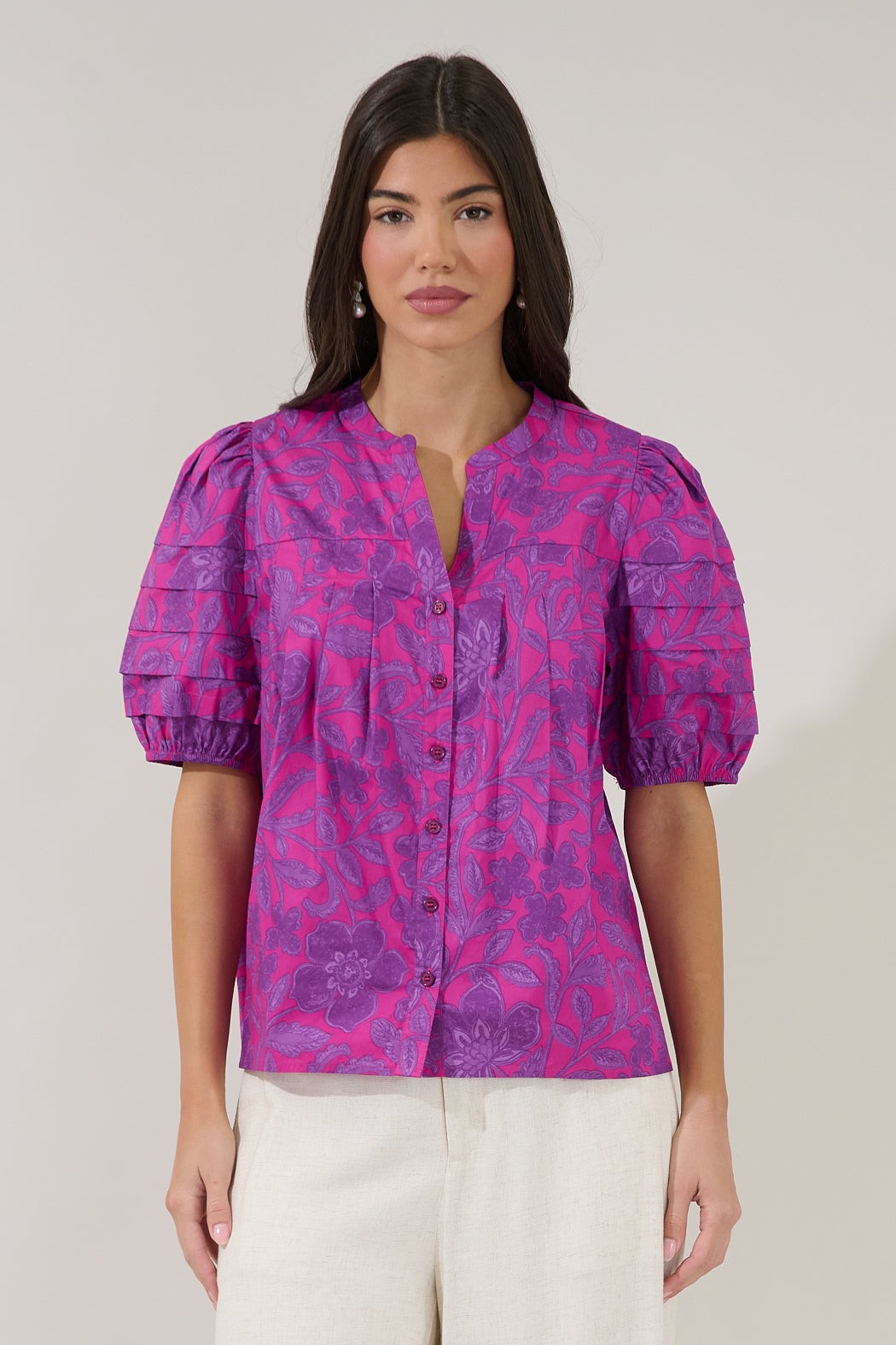 A woman modeling a short-sleeve button-front blouse with puff sleeves and a tonal floral jacquard pattern in bright pink and purple, paired with white pants against a plain light gray studio background.
