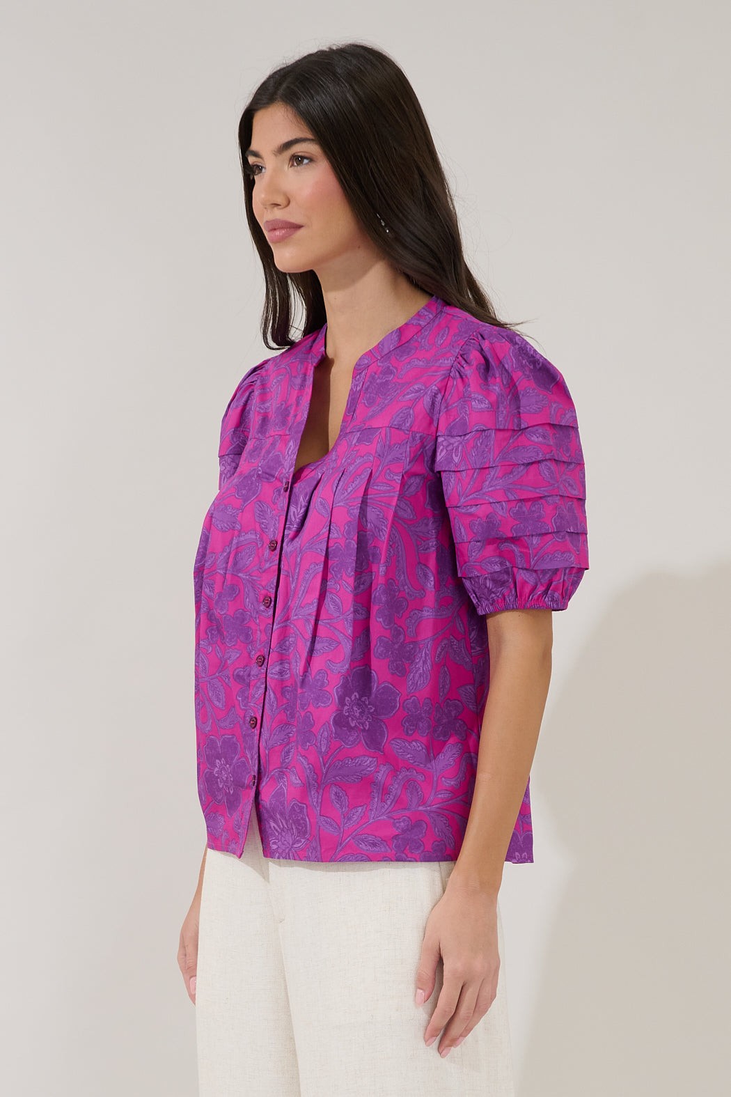 A woman wearing a short-sleeved button-up blouse with a vibrant magenta and purple abstract pattern, paired with white pants, photographed against a plain light beige studio background.