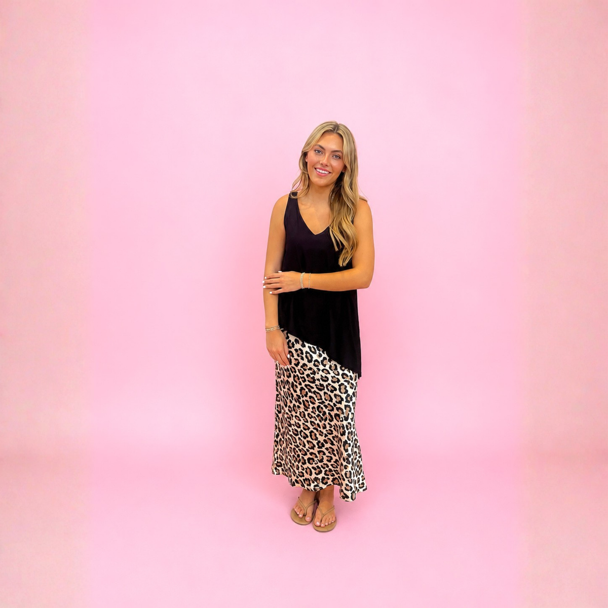 A woman modeling a two-piece outfit consisting of a black sleeveless top and a leopard print maxi skirt, standing against a solid pink background.