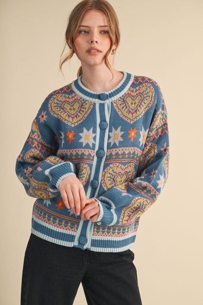 A woman wearing a blue Fair Isle knit cardigan sweater with heart and star patterns, paired with black pants, against a solid beige background.