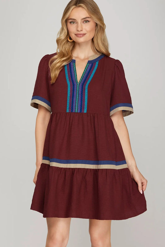 Woman wearing a burgundy tiered mini dress with puff sleeves and embroidered V-neckline in teal and blue, accented with taupe and navy trim on sleeves and hem.