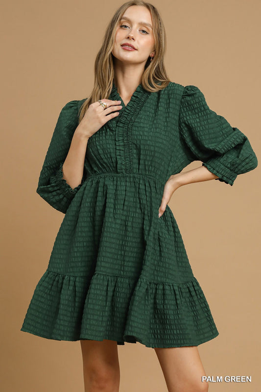 Woman wearing a textured tiered mini dress with ruffle-trim neckline and three-quarter puff sleeves, styled with soft waves and minimal jewelry.