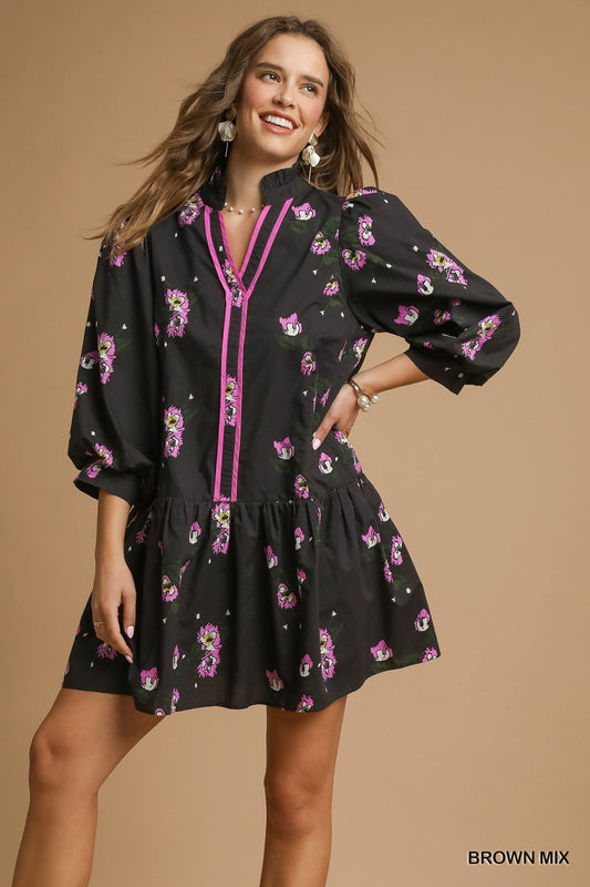 Woman wearing a black mini dress with pink floral print, puff sleeves, and hot pink contrast piping along a ruffled V-neckline and front placket.