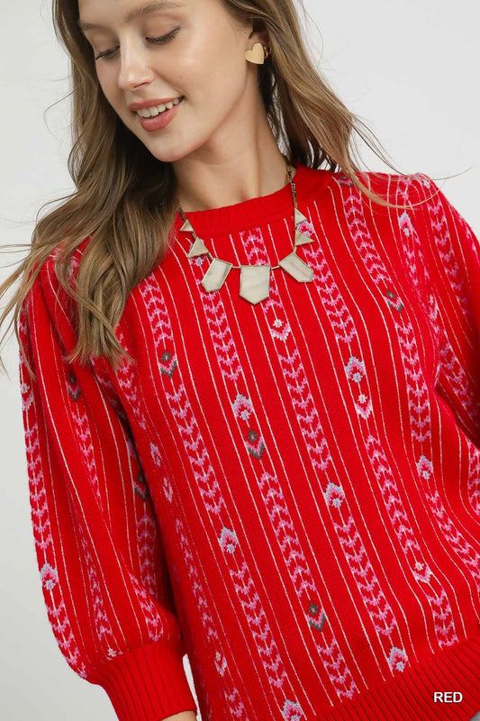 Woman wearing a red knit sweater with embroidered fair isle-inspired pattern, puff sleeves, and ribbed cuffs, styled with a statement necklace and heart-shaped earrings.
