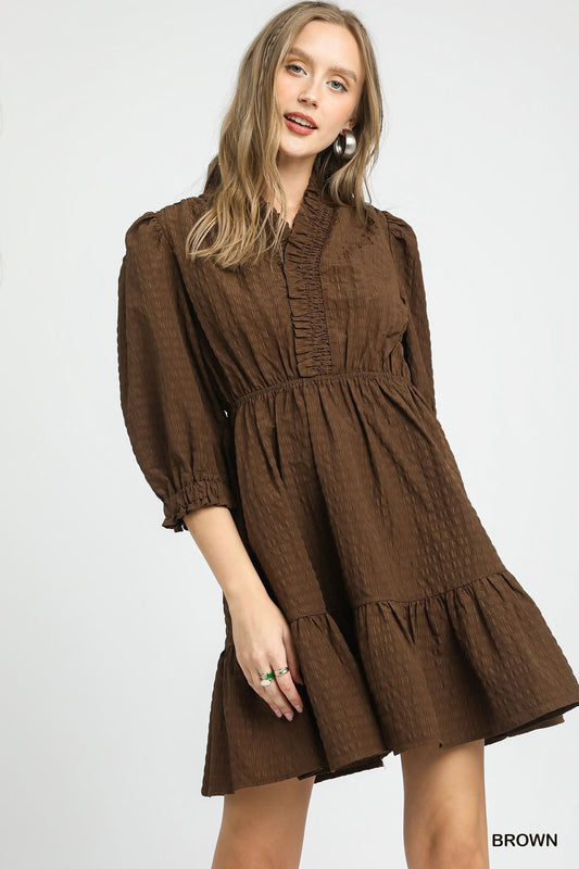 Model wearing a textured brown mini dress with a ruffle-trim V-neckline, puff sleeves, and a tiered skirt, styled with silver statement earrings.
