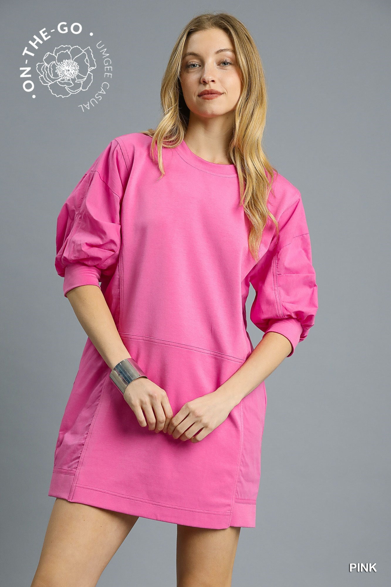 Model wearing a vibrant pink pullover sweatshirt dress with puff sleeves, relaxed fit, and sporty seaming detail, styled with a silver cuff bracelet and striped socks for a fun, casual look.