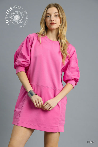 Model wearing a vibrant pink pullover sweatshirt dress with puff sleeves, relaxed fit, and sporty seaming detail, styled with a silver cuff bracelet and striped socks for a fun, casual look.