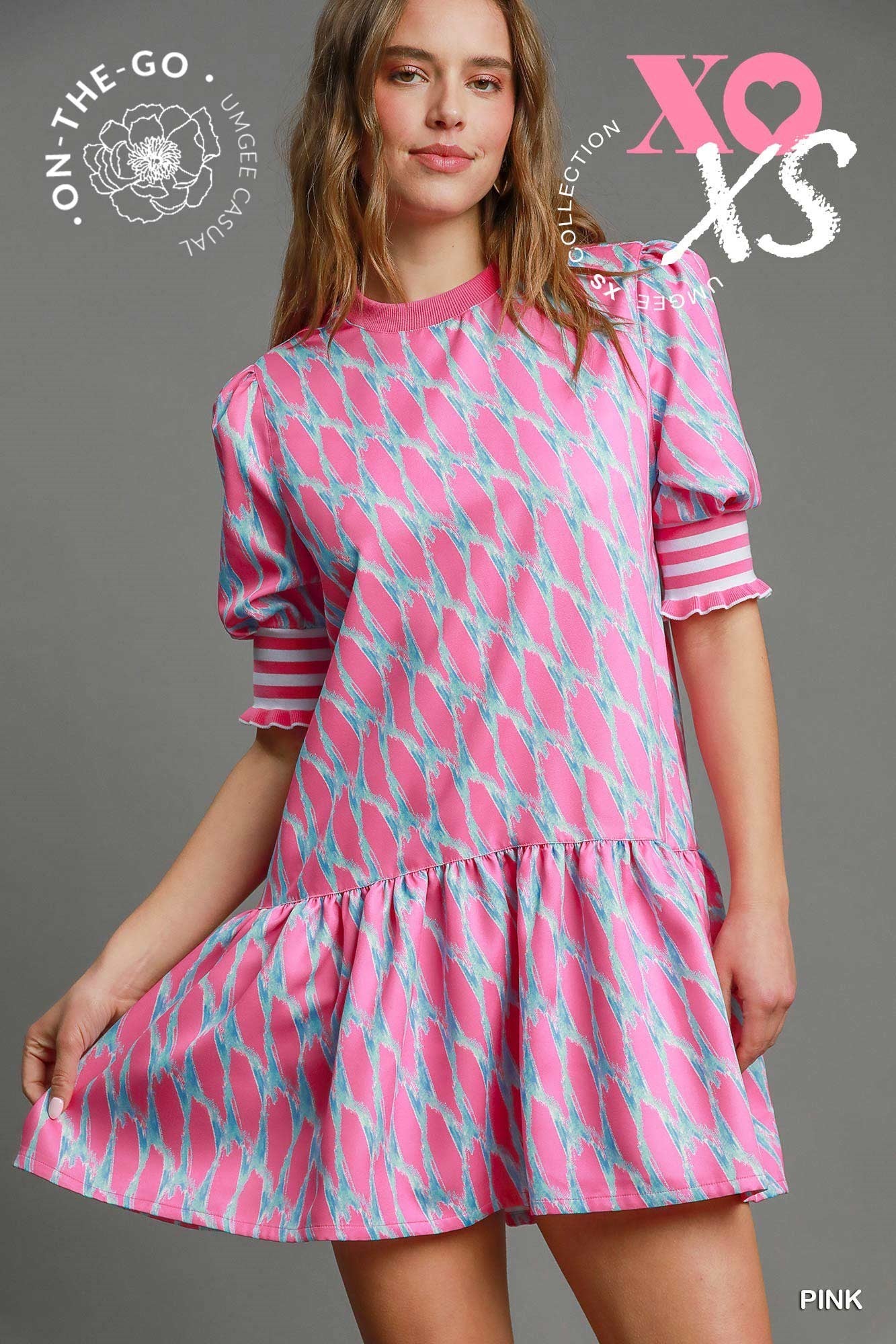 Woman wearing a pink and aqua printed mini dress with puff sleeves and striped cuffs, styled with blue flats, standing against a gray backdrop.