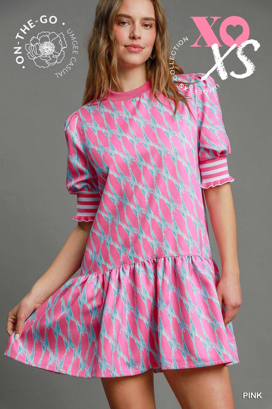 Woman wearing a pink and aqua printed mini dress with puff sleeves and striped cuffs, styled with blue flats, standing against a gray backdrop.