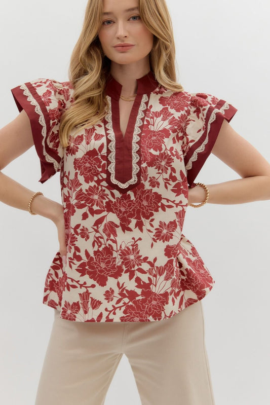 Model wearing a floral print blouse with flutter sleeves, lace trim, and a notched neckline, paired with light neutral trousers.