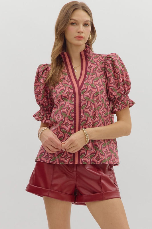 Woman wearing a pink and olive geometric print puff sleeve blouse with contrast trim V-neckline and smocked cuffs, styled with burgundy leather shorts – Milo & Lily Boutique.