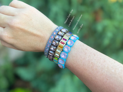 Woman's wrist wearing four colorful gemstone stretch bracelets labeled Maeve Dark Gray, Layla Brown, Penelope Rainbow, Griffin Blue