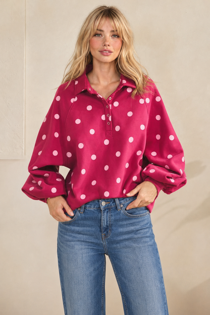 A woman modeling a bright pink long-sleeve button-up shirt with white polka dots, paired with blue jeans, against a neutral beige background.