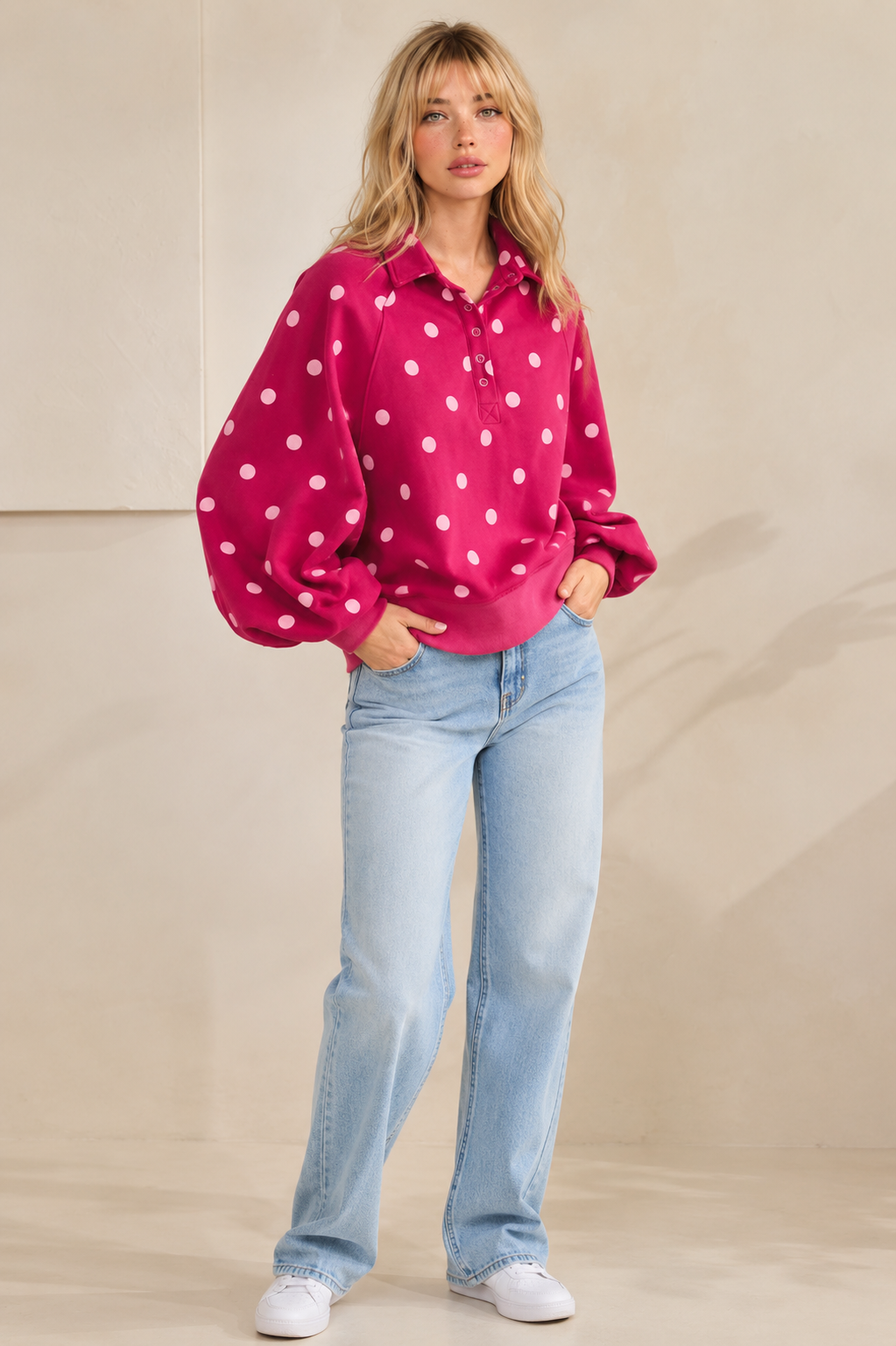A woman modeling a bright pink long-sleeve collared top with white polka dots, paired with light-wash blue jeans and white sneakers, standing against a neutral beige studio backdrop.