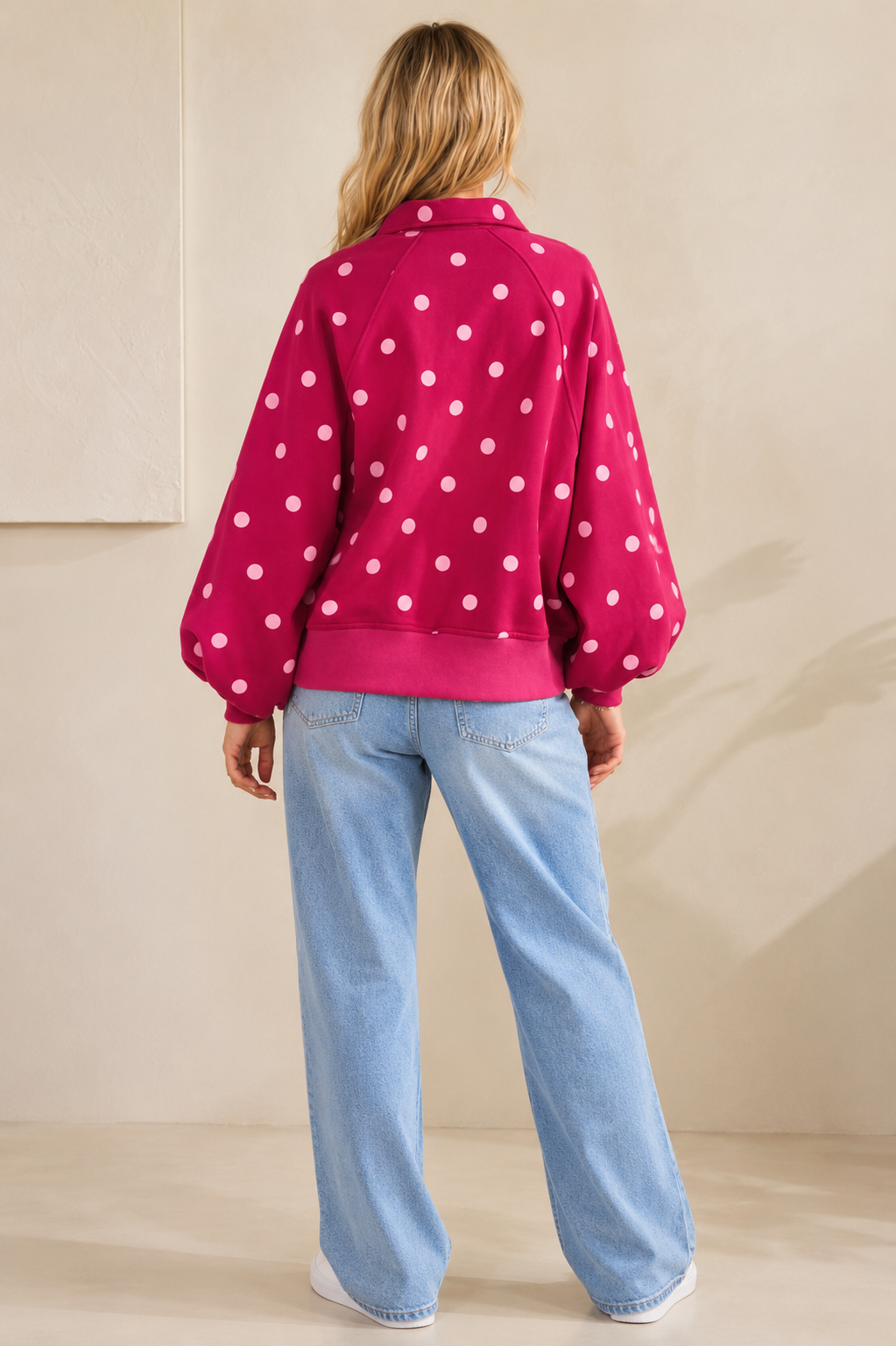 A woman shown from the back wearing a bright pink cropped sweatshirt with white polka dots and light blue jeans, standing against a neutral beige wall.