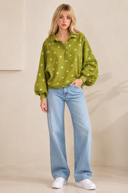 A woman modeling a green long-sleeve collared shirt with white polka dots, paired with light blue wide-leg jeans and white sneakers, standing against a beige studio wall.
