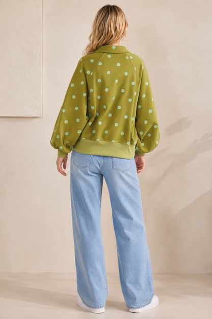 A woman viewed from behind wearing a light olive green long-sleeve sweatshirt with mint green polka dots, paired with light blue wide-leg jeans and white sneakers, set against a warm beige studio background.