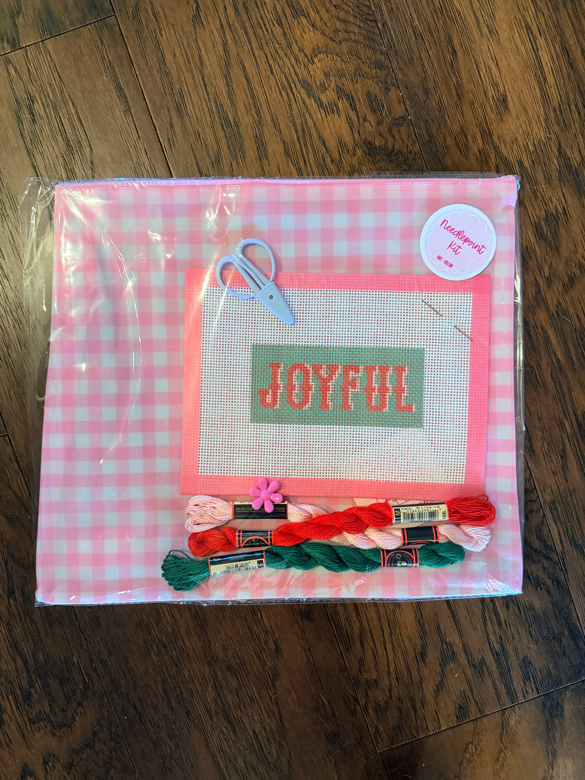 A packaged needlepoint kit on a dark wood floor. The kit features a pink and white gingham background, a central white canvas with a green rectangle containing the word 'JOYFUL' in red, pink border lines, a small blue-handled scissors, and several colorful threads (red, green, pink, white) and spools of thread at the bottom.