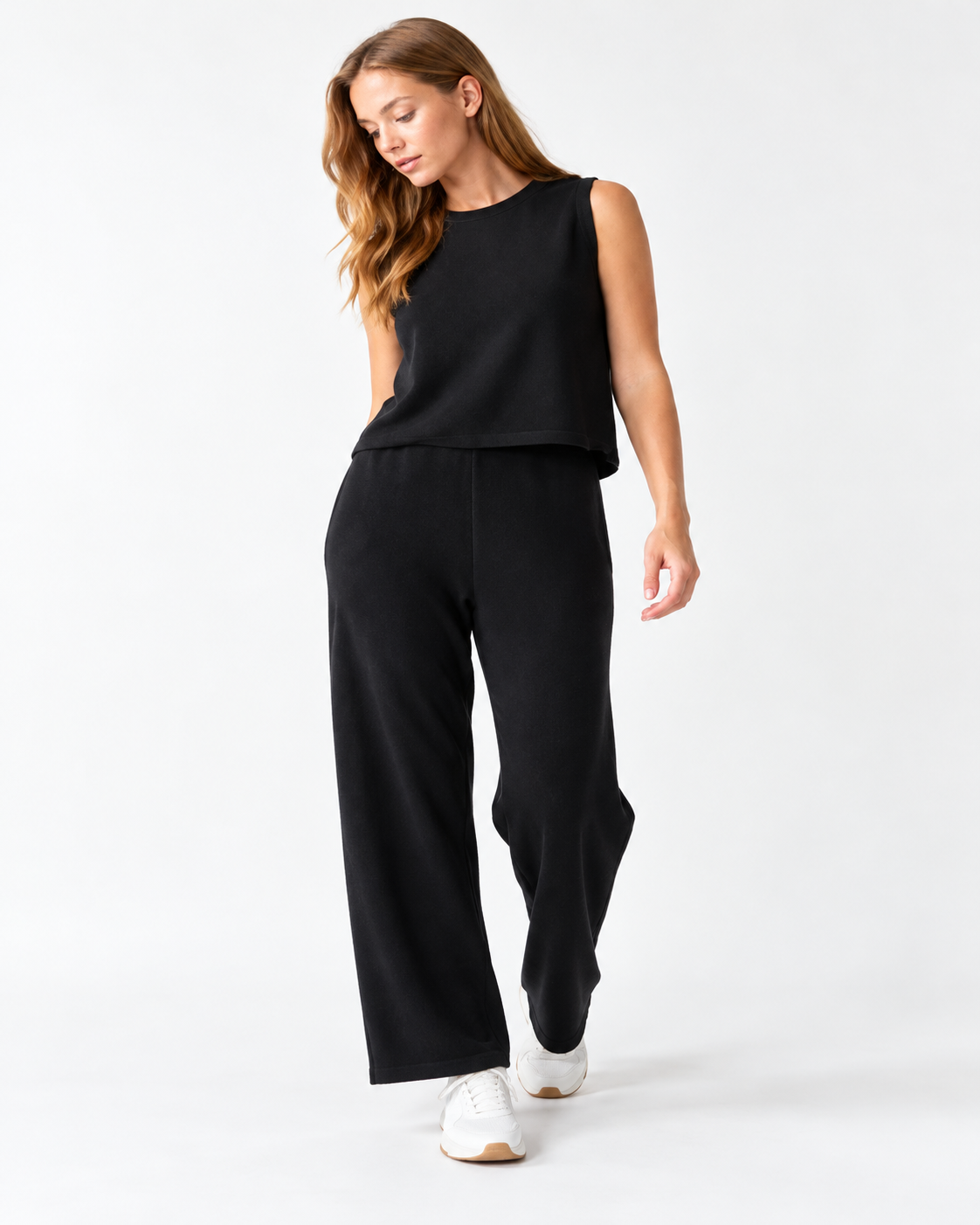 A woman modeling a black sleeveless top and matching wide-leg pants set, paired with white sneakers, against a plain white studio background.