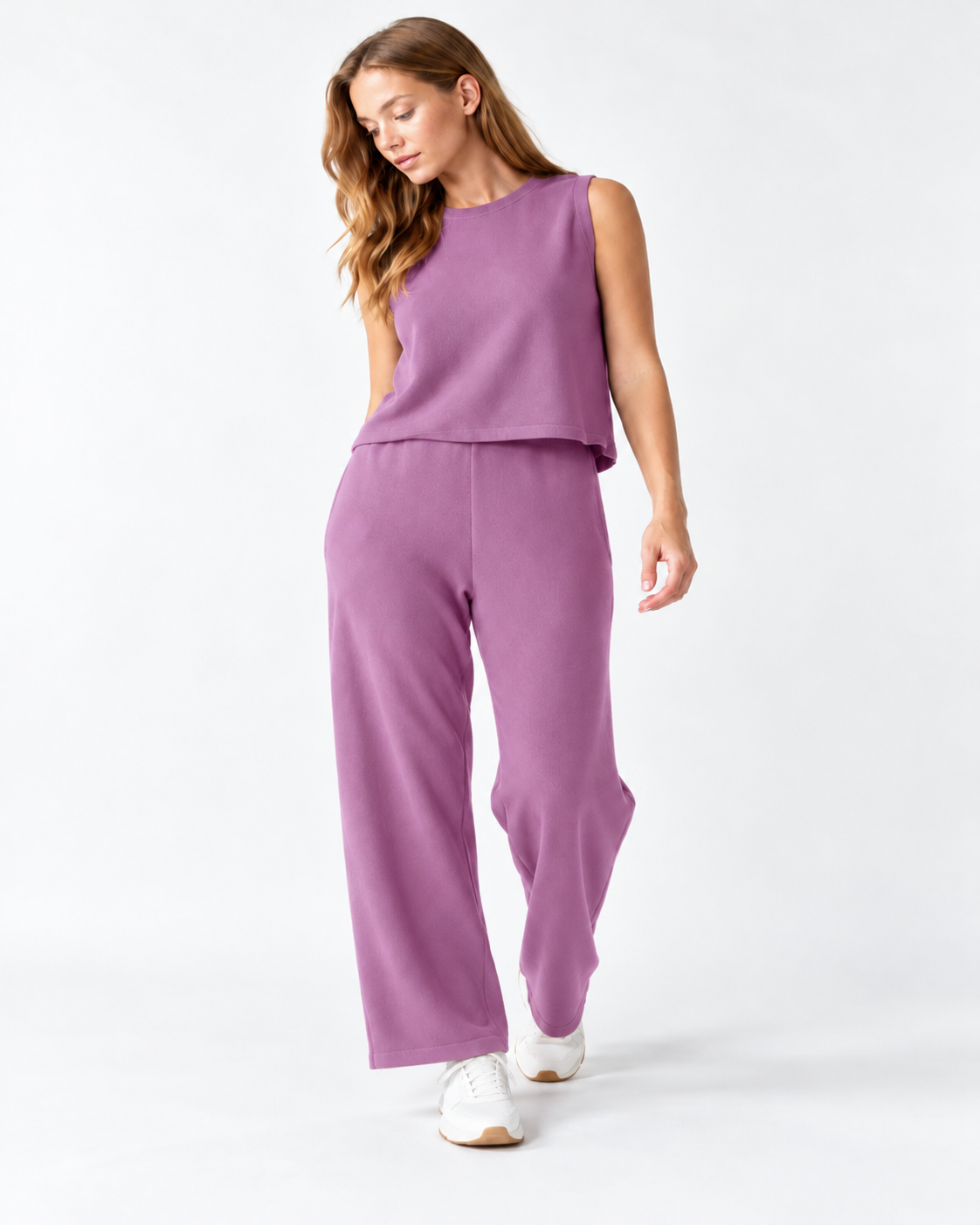 A woman modeling a matching two-piece lounge set in a dusty purple color, consisting of a sleeveless top and wide-leg pants, paired with white sneakers on a light gray studio background.