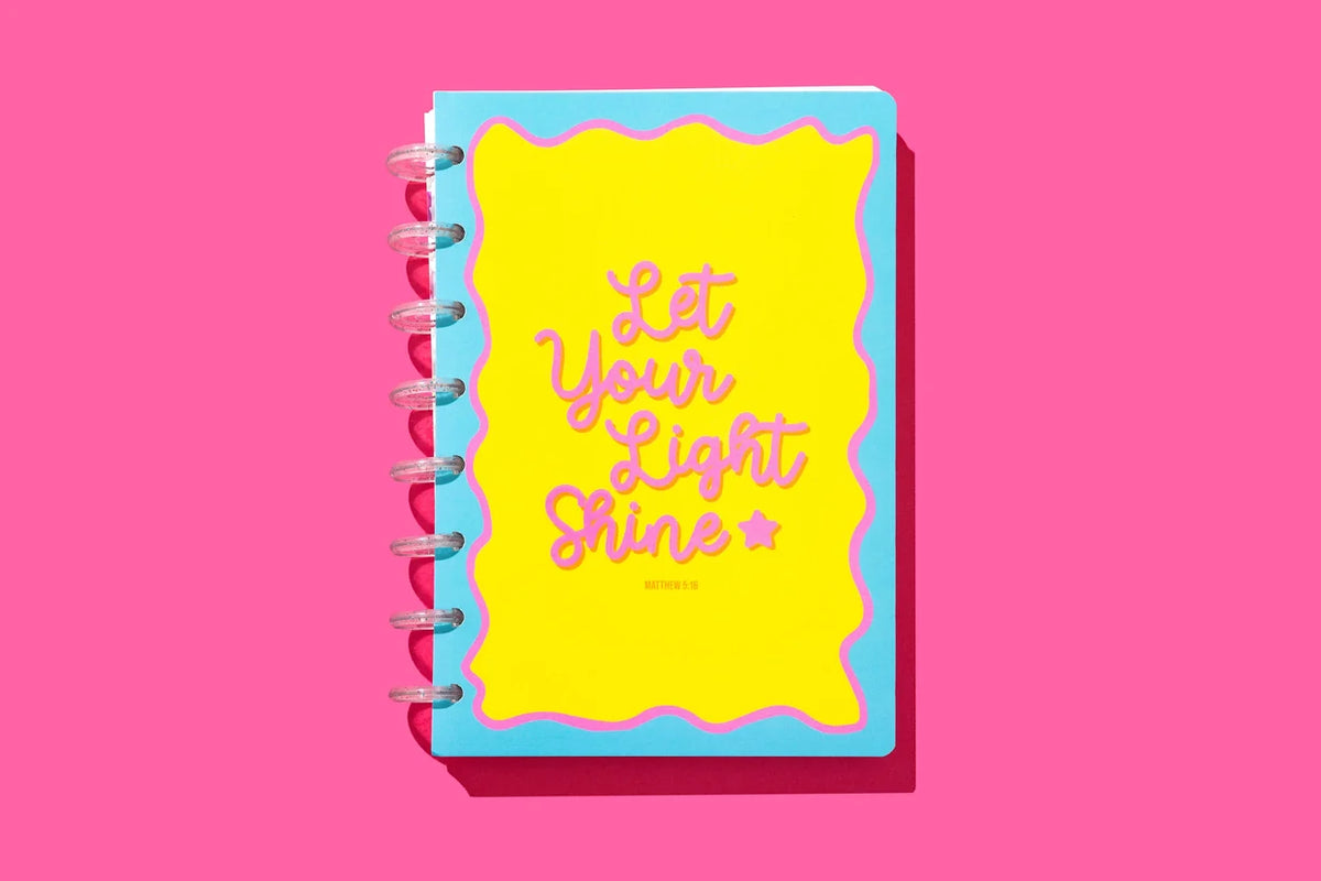 A spiral-bound notebook with a bright yellow cover and a light blue border, featuring the phrase 'Let Your Light Shine' in pink script. The notebook is shown on a solid pink background with a soft drop shadow.