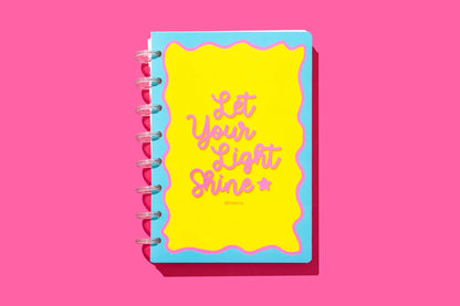 A spiral-bound notebook with a bright yellow cover and a light blue border, featuring the phrase 'Let Your Light Shine' in pink script. The notebook is shown on a solid pink background with a soft drop shadow.