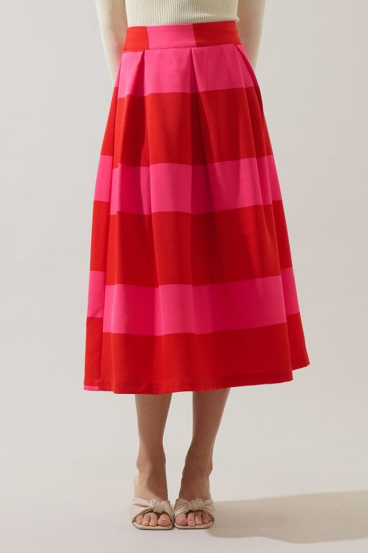 A model is shown from the waist down wearing a pleated midi skirt with bold horizontal stripes in red, orange, and pink, paired with beige flat sandals against a light gray studio background.