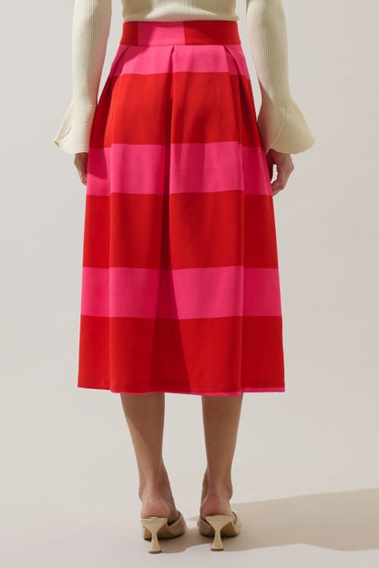A model from the back wearing a pleated midi skirt with bold horizontal stripes in red, hot pink, and coral, paired with a cream long-sleeve top and beige kitten heels.