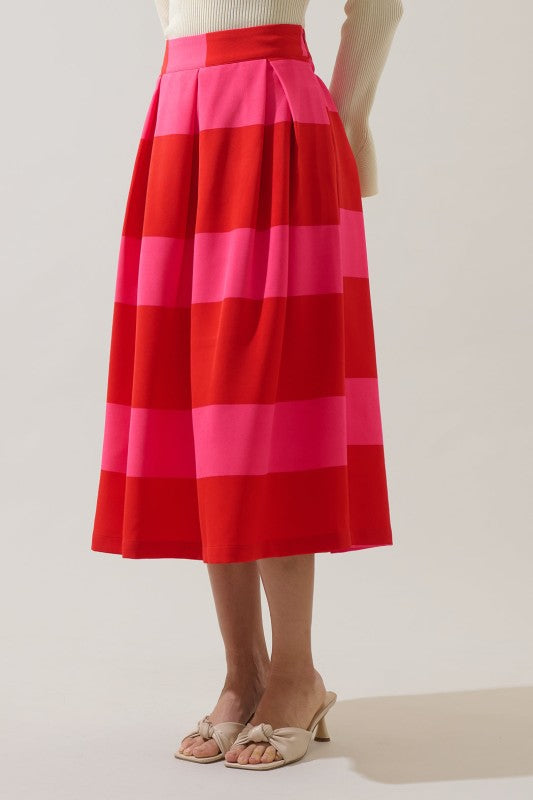 A model wearing a pleated midi skirt with bold horizontal stripes in red, pink, and orange, paired with beige heeled sandals against a plain studio background.