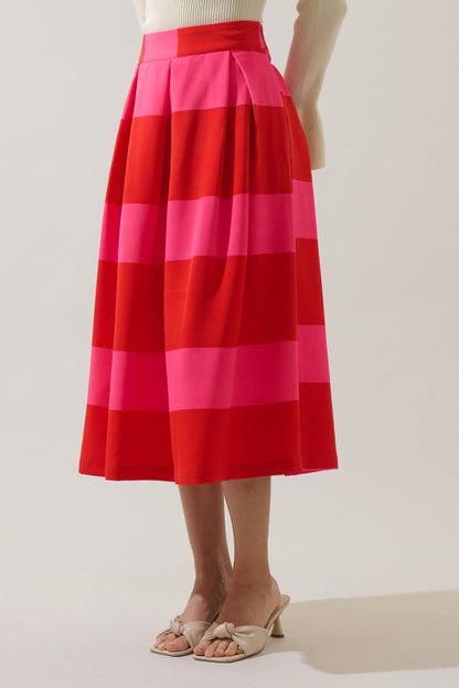 A model wearing a pleated midi skirt with bold horizontal stripes in red, pink, and orange, paired with beige heeled sandals against a plain studio background.
