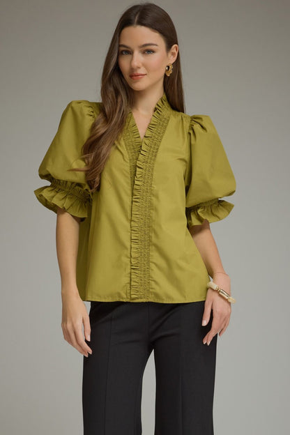 A woman modeling a sage green blouse with dramatic puff sleeves, ruffled trim, and a V-neckline, paired with black pants against a neutral gray studio background.