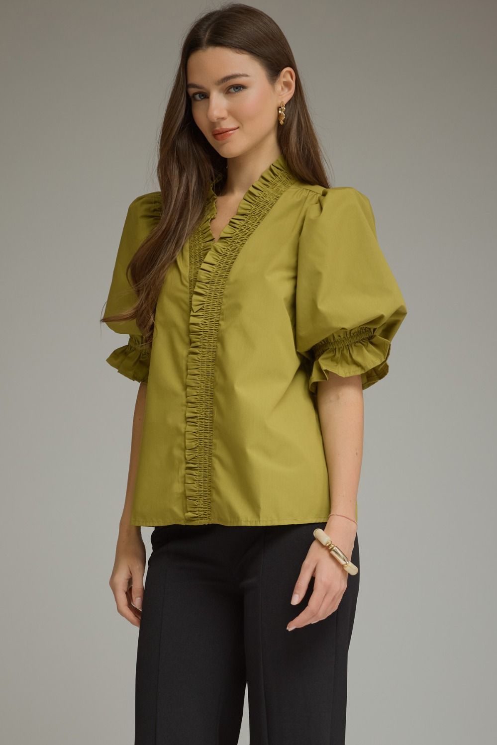 A woman modeling a short-sleeved olive green blouse with ruffled trim along the V-neckline and cuffs, paired with black pants, against a solid gray studio background.