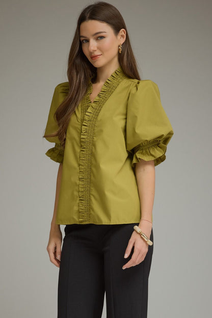 A woman modeling a short-sleeved olive green blouse with ruffled trim along the V-neckline and cuffs, paired with black pants, against a solid gray studio background.