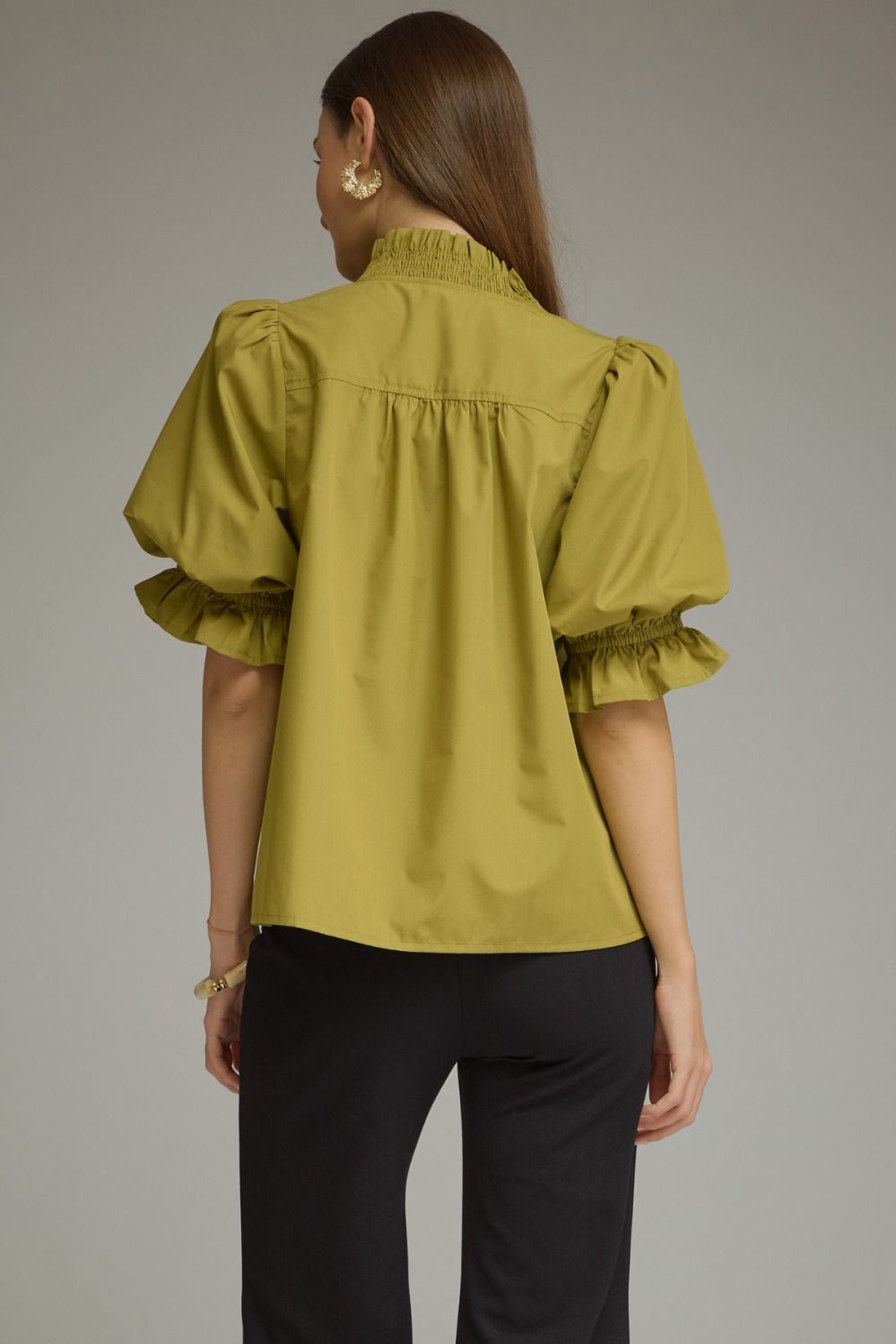 A woman shown from the back wearing a mustard yellow blouse with puffed short sleeves and a smocked neckline, paired with black pants, against a neutral gray studio background.