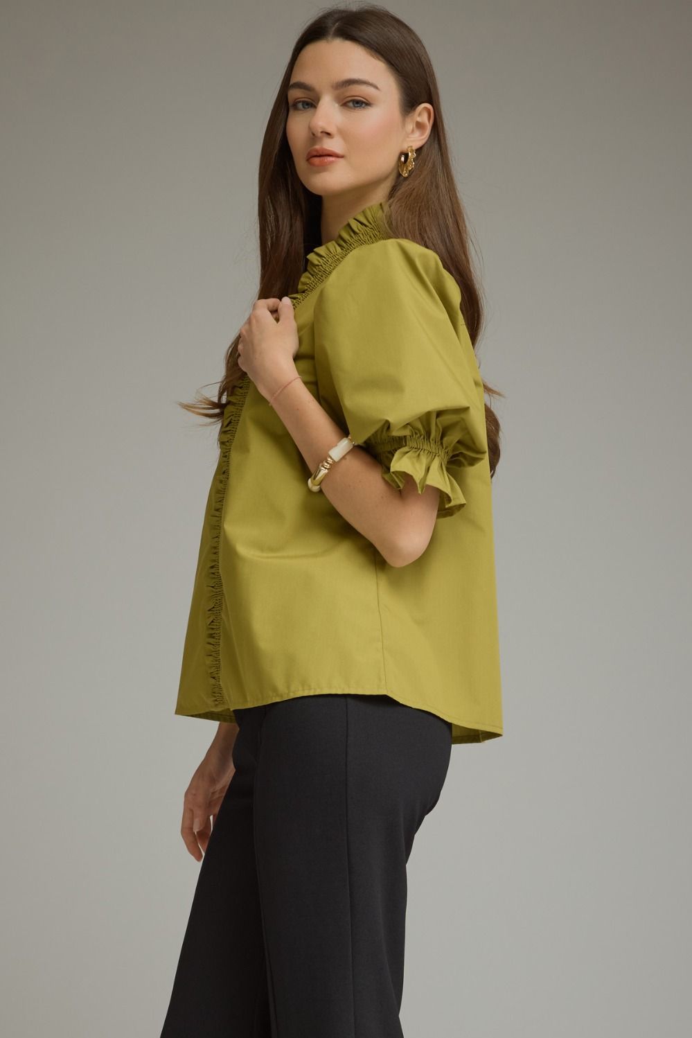 A woman modeling a short-sleeved olive green blouse with ruffled trim and puff sleeves, paired with black trousers, against a plain gray studio background.