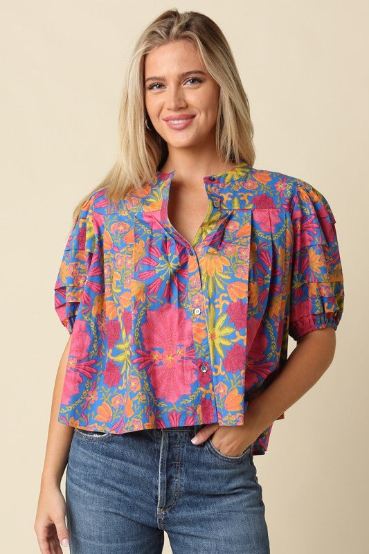 A woman wearing a colorful short-sleeve button-up blouse with a vibrant floral and paisley print in pink, blue, yellow, and orange, paired with blue jeans, set against a solid beige background.