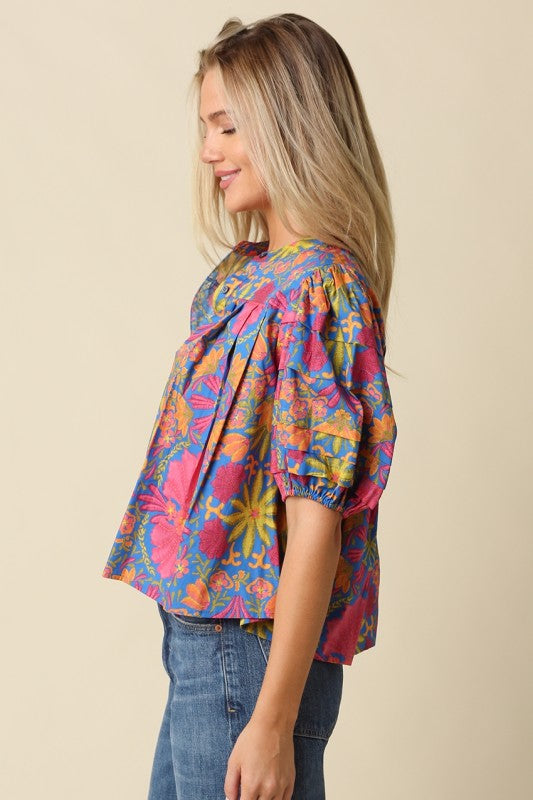 A woman wearing a colorful, short-sleeved blouse with a vibrant abstract pattern in pink, blue, yellow, and orange, paired with blue jeans, photographed against a solid beige background.