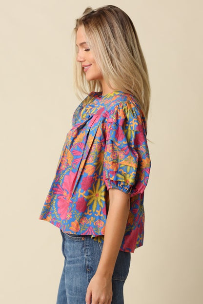 A woman wearing a colorful, short-sleeved blouse with a vibrant abstract pattern in pink, blue, yellow, and orange, paired with blue jeans, photographed against a solid beige background.