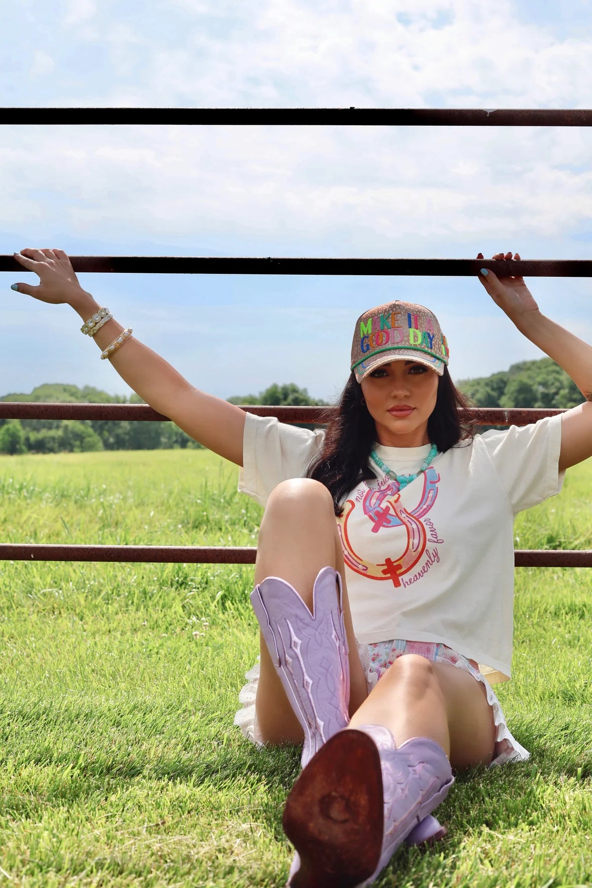A woman sitting on a grassy field behind a metal fence, wearing a white graphic t-shirt, a colorful baseball cap, and light purple cowboy boots.
