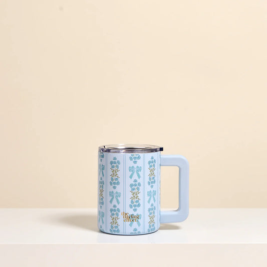 A light blue handled mug with a vertical pattern of bows and flowers in blue and gold, featuring a stainless steel rim and a matching light blue handle, set against a warm beige background.